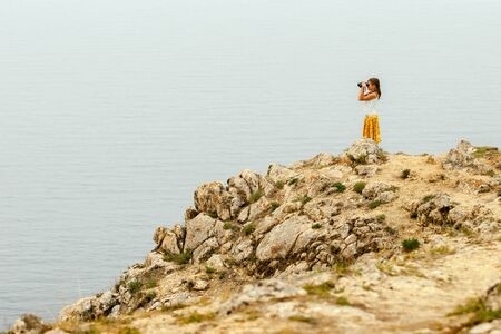 A child traveler girl stands on a rock above the water of Lake Baikal and looks into the distance with binoculars. Big stones. T-shirt and colored yellow skirt.の写真素材