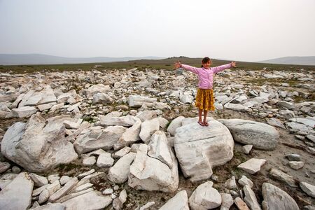 Girl child stands with arms outstretched on a large white stone in the steppe. In a purple jacket and a yellow skirt. Misty sky and hills from behind. A lot of big white stones. Copy space.の写真素材