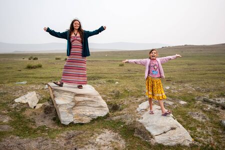 Cheerful smiling mother and daughter are standing on large white stones in the steppe with arms outstretched. Child with his tongue hanging out. Morning with fog. Hills on the horizon. Green grass. Copy space.の写真素材
