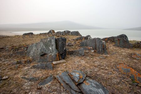 Beautiful black rocks sticking out of the ground with a lake and hills, foggy morning. Stones with red moss like rust. Dead grass. Grey sky. Copy space.の写真素材