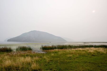 Lake in the fog and a fisherman in a boat with green grass near the shore. Peace and silence. Big rock in the water. The faded sun shines through the fog. Grey sky. Copy space.の写真素材