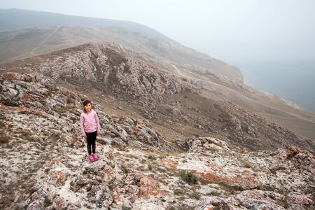 A young girl tourist stands on the edge of a high cliff on the shore of Lake Baikal. Girl looking at the camera. The cliffs are red covered with moss. The weather is foggy.の写真素材