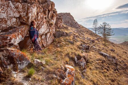 A girl in a colored dress and shawl stands by a beautiful rocky ridge covered with red moss on the side of a mountain. European face with long black hair. Copy space.の写真素材