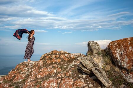 A beautiful girl in a dress with a waving scarf stands on a rock against a blue sky with clouds. European appearance, black hair. The stones on the rock are covered with red moss. There are clouds in the sky. Copy space.の写真素材