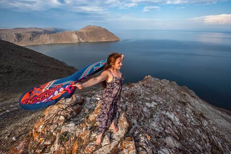 A cute young girl in a dress with a colorful scarf fluttering behind stands on a rock above Lake Baikal. European appearance, long black hair. The water is calm. Beautiful rock. Copy space.の写真素材