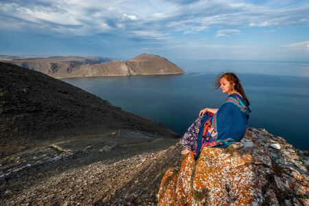 A nice girl in a dress with a colorful scarf on her shoulders sits on a rock by Lake Baikal. European appearance, long black hair. The water is calm. Beautiful rock and sky with clouds. Copy space.の写真素材