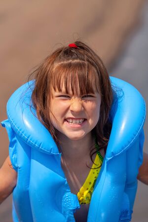Child girl in a blue inflatable life jacket makes a grimace. The eyes are narrow, half-closed. The background is blurry. Copy space. Vertical.の写真素材
