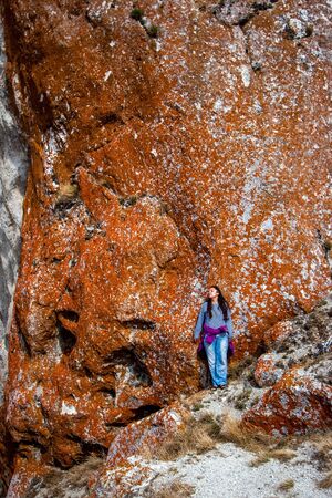 A girl stands by a huge stone wall with red moss. The eyes are half closed. Head turned to the side. Long black hair, dressed in blue pants and a striped sweater.の写真素材