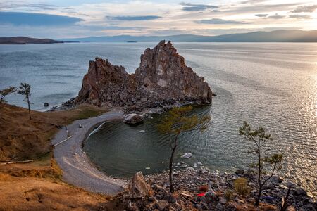View of the granite rock standing standing on the cape. On the shore of stones and grass. Ripples on the lake water. Mountains beyond the lake. Sunset. There are clouds in the sky.の写真素材