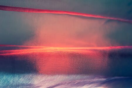 Abstract blurred background. Very colorful thin airy beautiful fabric with tints of red and blue. Texture of tulle in the sun at sunset. Shallow depth of field.の写真素材