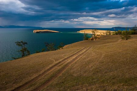 View of the island on Lake Baikal across the steppe with a field road in cloudy weather with the sun. Trees grow on the edge of the shore, on the horizon there are mountains. Heavy clouds in the sky.の写真素材