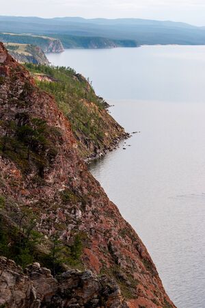 The red steep cliffs of Olkhon Island on Lake Baikal stretching into the distance. On the rocks grow trees and moss. Grey sky.の写真素材