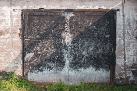 Old grunge garage door with black iron doors and iron door hinges. The gates are all scratched and scribbled. Around the plastered wall. Bottom green grass.の写真素材