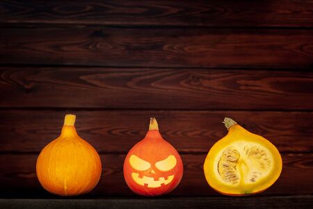 Three pumpkins lie next to a wooden brown background. One red pumpkin with halloween face and two yellow. One pumpkin is cut and seeds are visible inside. Copy space.の写真素材
