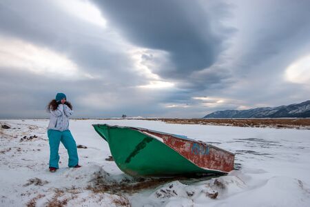 A girl pulling a hat over her ears stands next to a boat by the river with ice in the winter against a beautiful sky. Girl with long dark hair. Mountains on the horizon.の写真素材