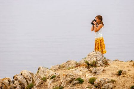 A girl with binoculars in her hands stands on Lake Baikal on the rocks and looks over the...