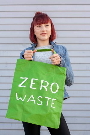 Green eco bag with a white inscription zero waste held by a young girl with red hair. Smiling girl in a denim jacket on a striped gray background.の写真素材