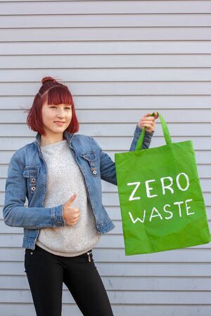 Girl with red hair holds a green textile eco bag with a white inscription zero waste and shows a thumb up. The girl is wearing a denim jacket and pants. Grey background. Copy space. Vertical.の写真素材
