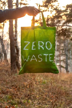 Zero waste concept. Textile green bag with a white inscription in hand on a background of nature and the sun. Against the background of blurry trees and autumn grass. Vertical. Copy space below.の写真素材