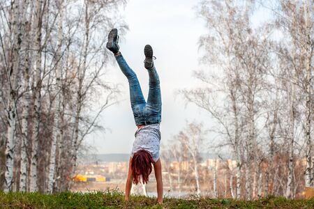 A girl with red hair stands on her hands with her back to the camera on a hill against the...