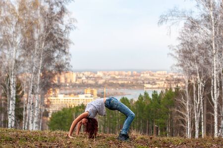 A young girl makes an acrobatic bridge on nature against the backdrop of a blurred city and forest....