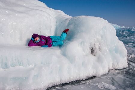 A girl in winter clothes lies on a huge ice sofa on a sunny day on Lake Baikal. Smooth ice glistens...