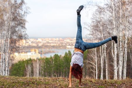 A girl with red hair makes an acrobatic wheel on nature against the background of a blurred city and forest. The girl has red hair, jeans. Motion blur. Grass on the ground. Acrobatics in nature.の写真素材