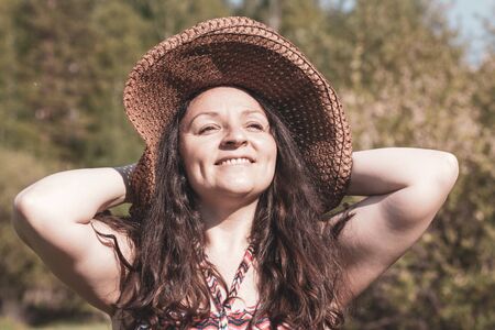 The girl in a straw hat smiles with his hands behind his head. Retro style. Sunny. Blurred background. Color tinting. Horizontal.の写真素材