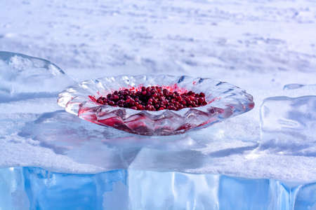 Red cranberries in a plate of ice stands on a piece of ice. The ice is sprinkled with snow. Russian Siberian tradition of meeting guests. Focus on a plate with a berry. Horizontal.の写真素材