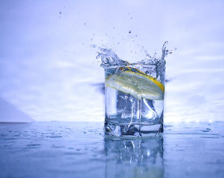 Glass of cold clear splashing water with falling lemon's slice on light-blue background