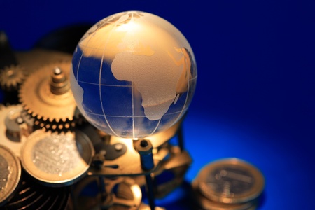 Glass globe lying on gears and coins on dark blue background