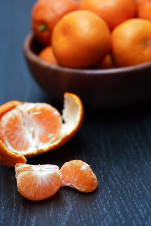 Peeled tangerine near wooden bowl full of fruits on black tableの写真素材