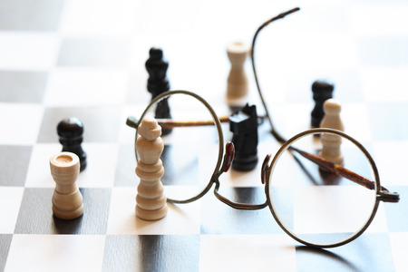 Black and white chessman set near spectacles on chessboard against lightの写真素材