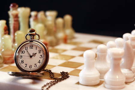 Set of chess pieces made from Onyx near watch on board against dark backgroundの写真素材
