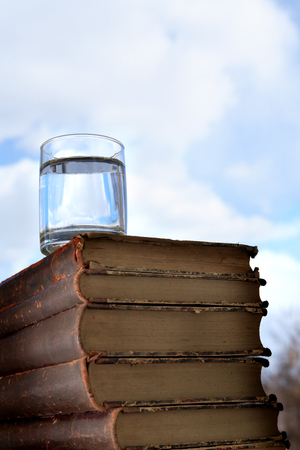 Glass of water on stack of old books on blue sky background with free spaceの写真素材