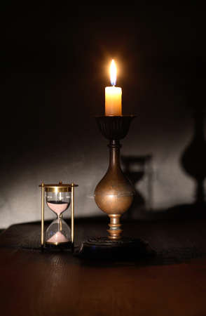 Elegant brass candlestick with lighting candle near hourglass on dark backgroundの写真素材