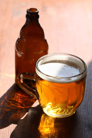 Mug of beer with foam on wooden table under sunlightの写真素材