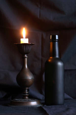 Glass black bottle near lighting candle against nice dark textile backgroundの写真素材