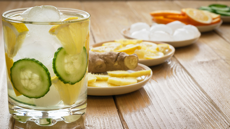Cold water with ice, ginger root, lemon and cucumber. Ingredients for making a refreshing drink on a wooden table. Healthy lifestyle.の写真素材