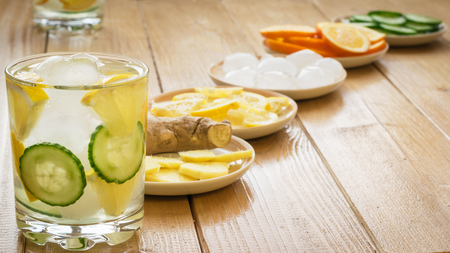 Cold water with ice, ginger root, lemon and cucumber. Ingredients for making a refreshing drink on a wooden table. Healthy lifestyle.の写真素材