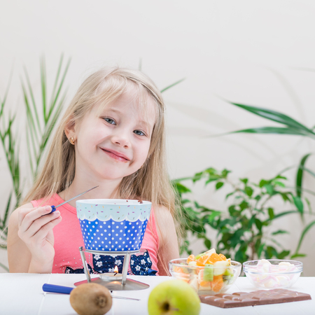 Little girl preparing and eating a chocolate fondue. Chocolate fondue with kiwi, banana, apple, tangerine and marshmallow. Happy little girl.の写真素材