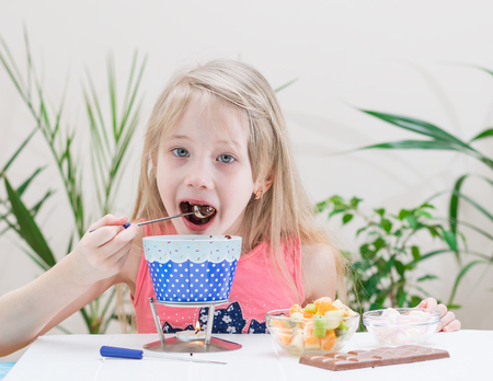 Little girl preparing and eating a chocolate fondue. Chocolate fondue with kiwi, banana, apple, tangerine and marshmallow. Happy little girl.の写真素材