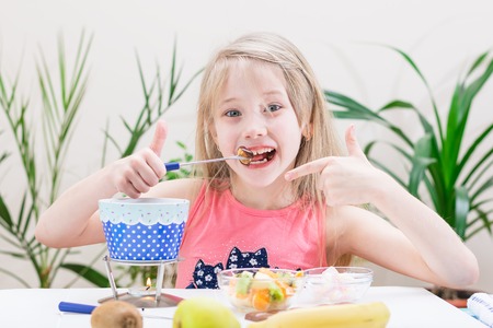 Little girl preparing and eating a chocolate fondue. Chocolate fondue with kiwi, banana, apple, tangerine and marshmallow. Happy little girl.の写真素材