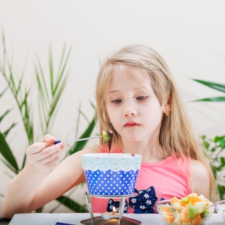 Little girl preparing and eating a chocolate fondue. Chocolate fondue with kiwi, banana, apple, tangerine and marshmallow. Happy little girl.の写真素材