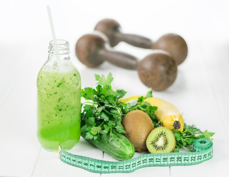 A bottle of cucumber smoothies, fruit and measuring tape on a white table. The dumbbells in the background. The concept of sports diet food for weight control.の写真素材