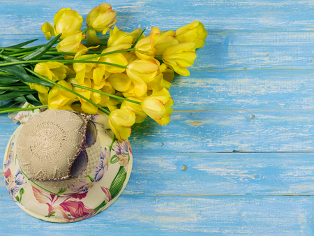 Hat with flowers and sunglasses on a blue wooden table. Hat with a bouquet of tulips on a wooden table.の写真素材