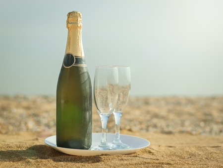 A bottle of sparkling wine with glasses on a white plate. Sandy summer beach in the sun.の写真素材
