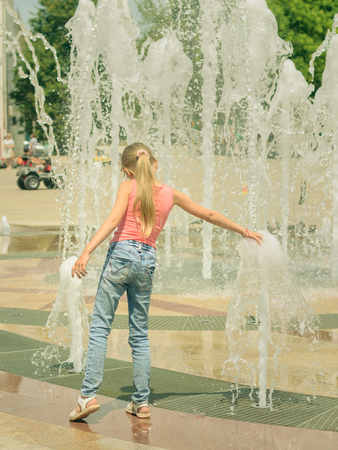 Girl in jeans plays with the water jets in the fountain. A child plays with water on a hot summer day.の写真素材