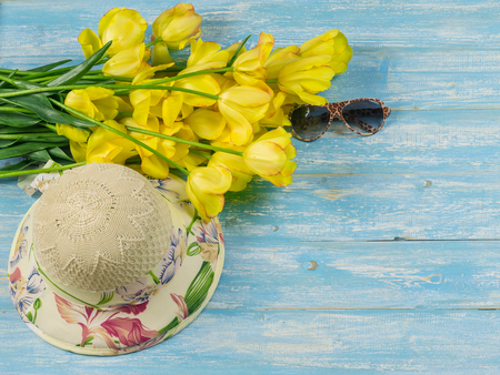 Hat with flowers and sunglasses on a blue wooden table. Hat with a bouquet of tulips on a wooden table. The concept accessories for a summer vacation.の写真素材