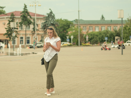 Young beautiful woman in the town square communicates through an electronic device. Portrait of woman in full growth in the city.の写真素材